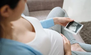 Mujer embarazada sentada en un sofá tocando su vientre mientras observa una fotografía de ecografía.