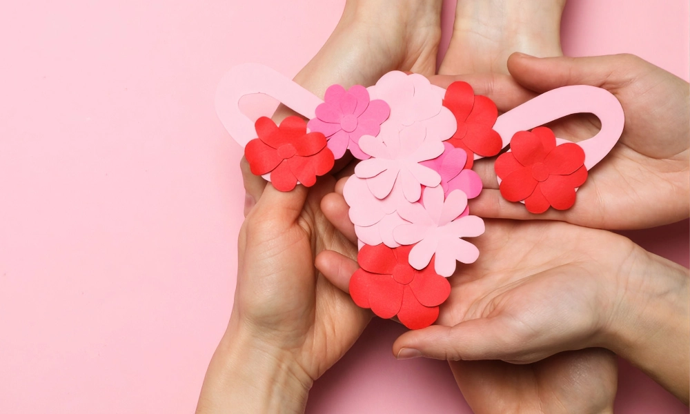 Múltiples pares de manos sostienen una representación artística del aparato reproductor femenino hecha con recortes de papel rosa y flores rojas y rosadas sobre un fondo rosa.